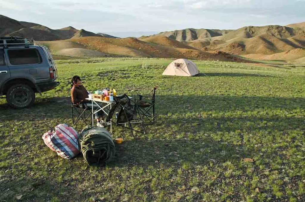 Our camp site for the first night on this trip to the Mongolian Gobi Altai. My cook sits at our dinner table. The dry creek bed is off to the right, my swag is in the foreground, and the only vegetation in sight is a very low green herb.